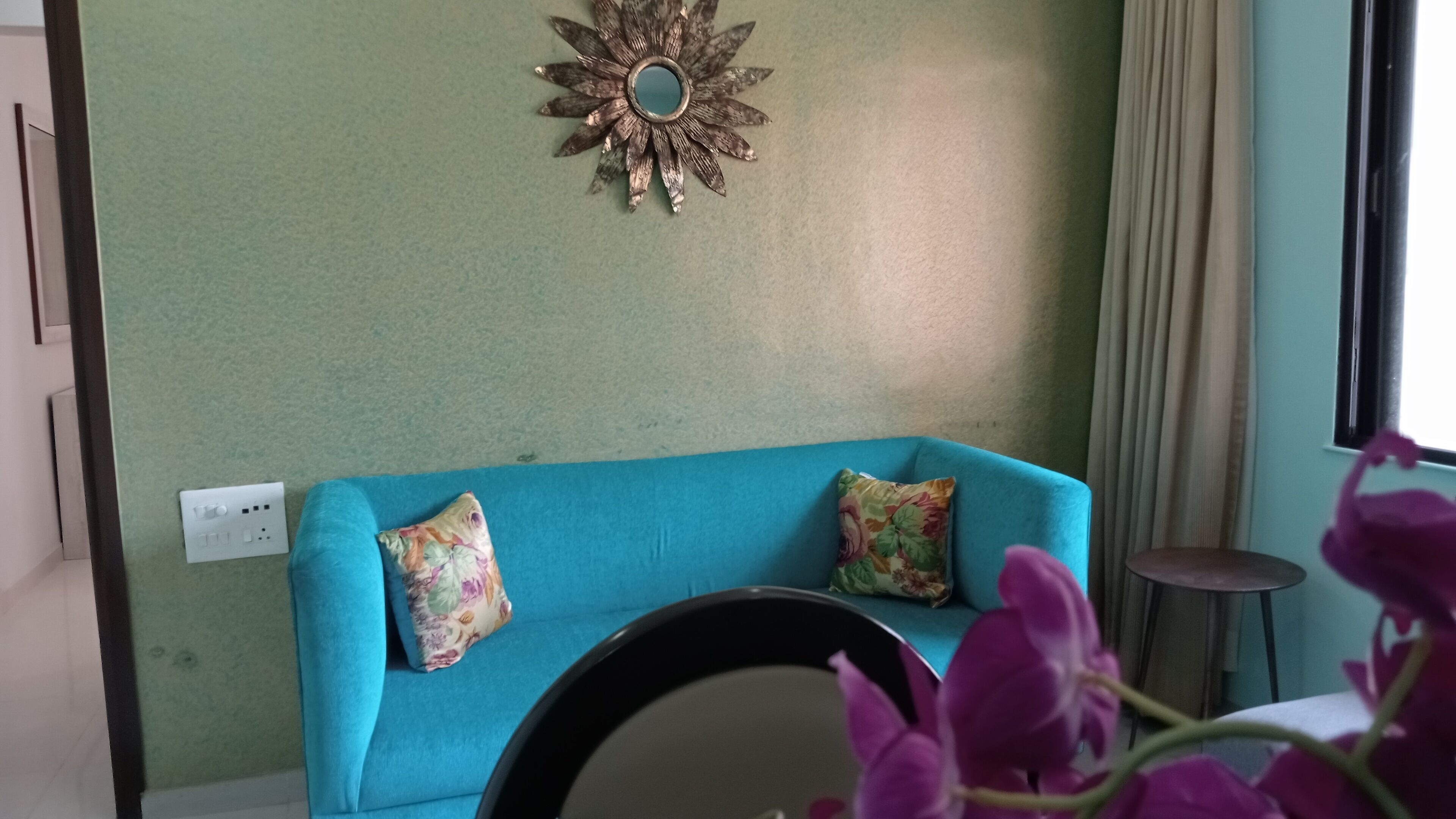 A room with a blue sofa, two floral cushions, a wall-mounted decorative mirror, and a window with curtains.
