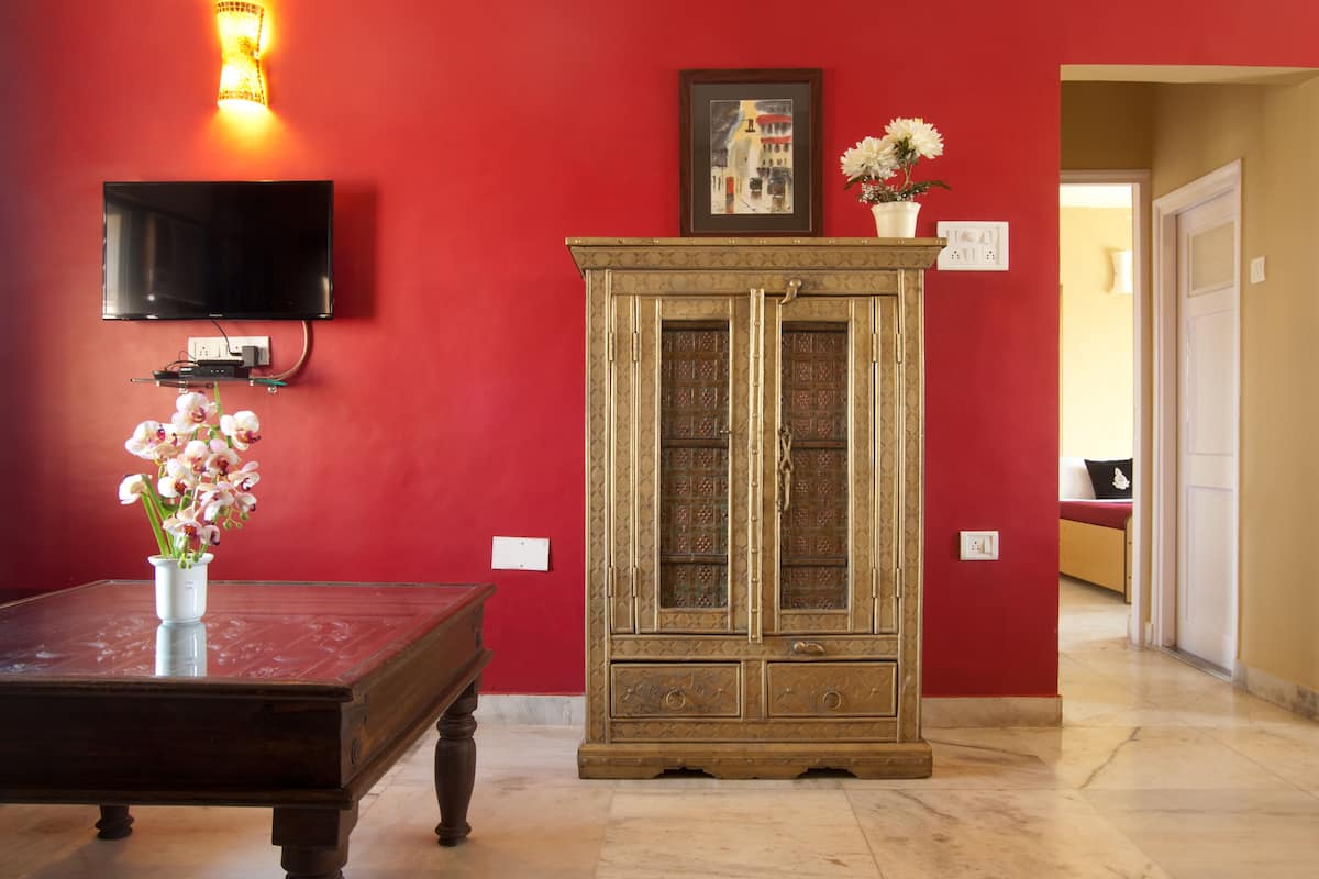 A room with a red wall, a TV mounted above a wooden table with a vase of flowers, and a cabinet with glass doors.