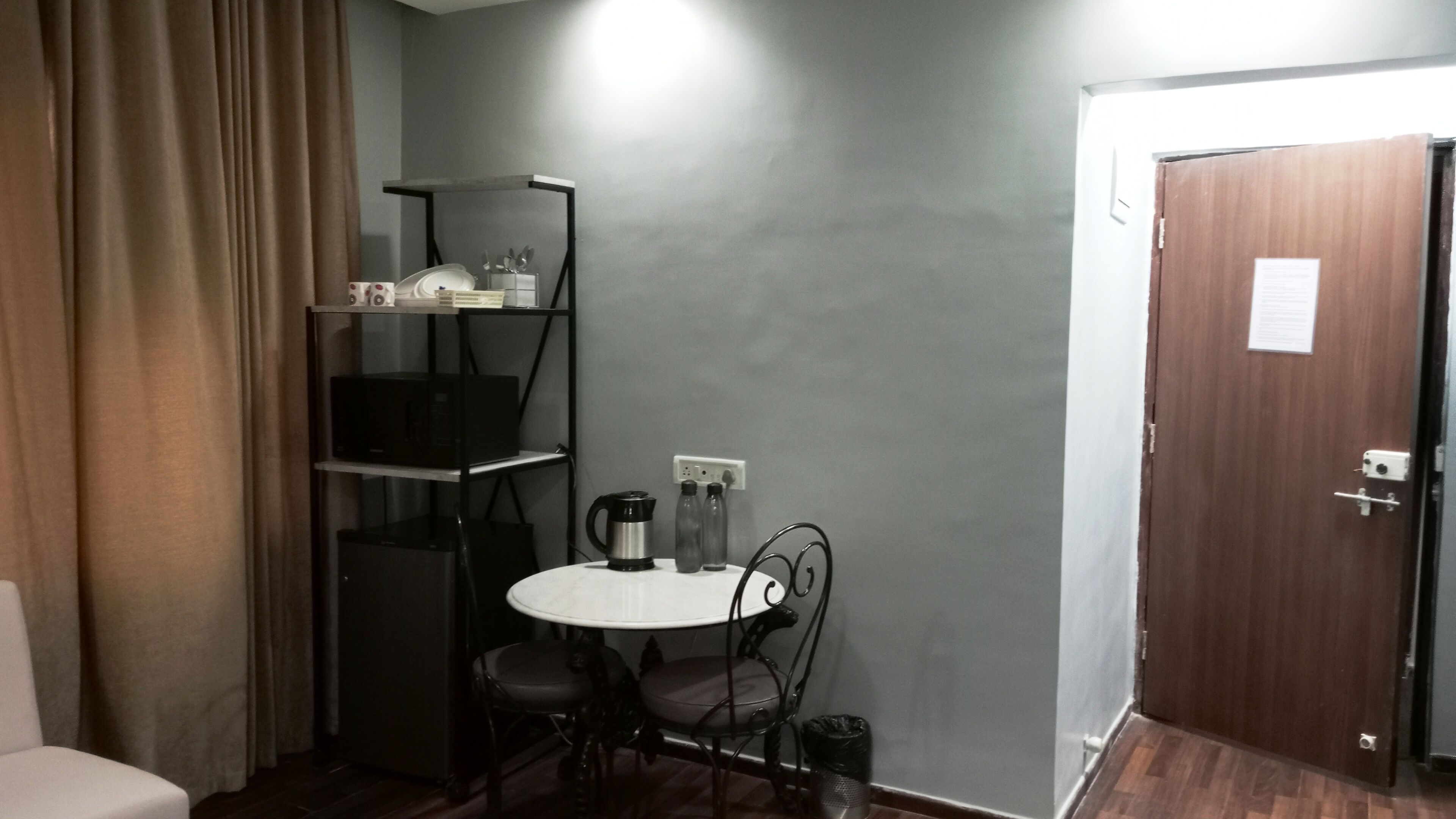 A small room with a single table and two chairs, a shelf with various items, a door, and a curtain.
