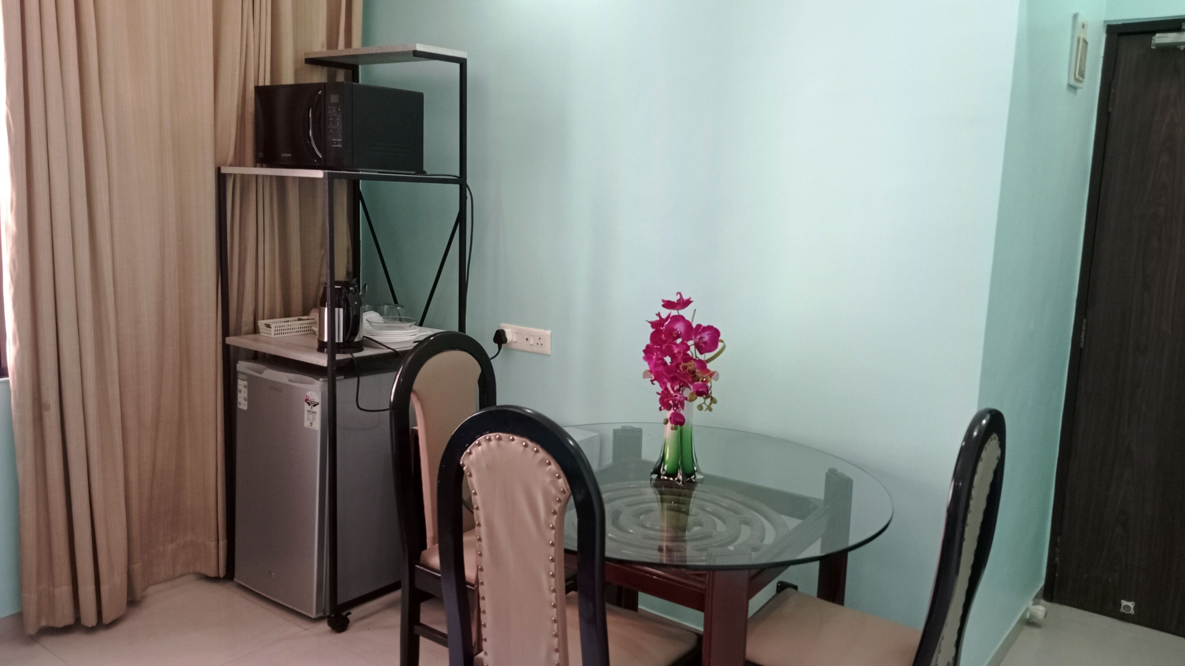 A compact room with a glass table, two chairs, a small refrigerator, a shelf unit, and a vase with flowers.