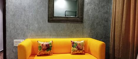 Una habitación con sofá amarillo, dos cojines florales, un espejo y una ventana con cortinas.