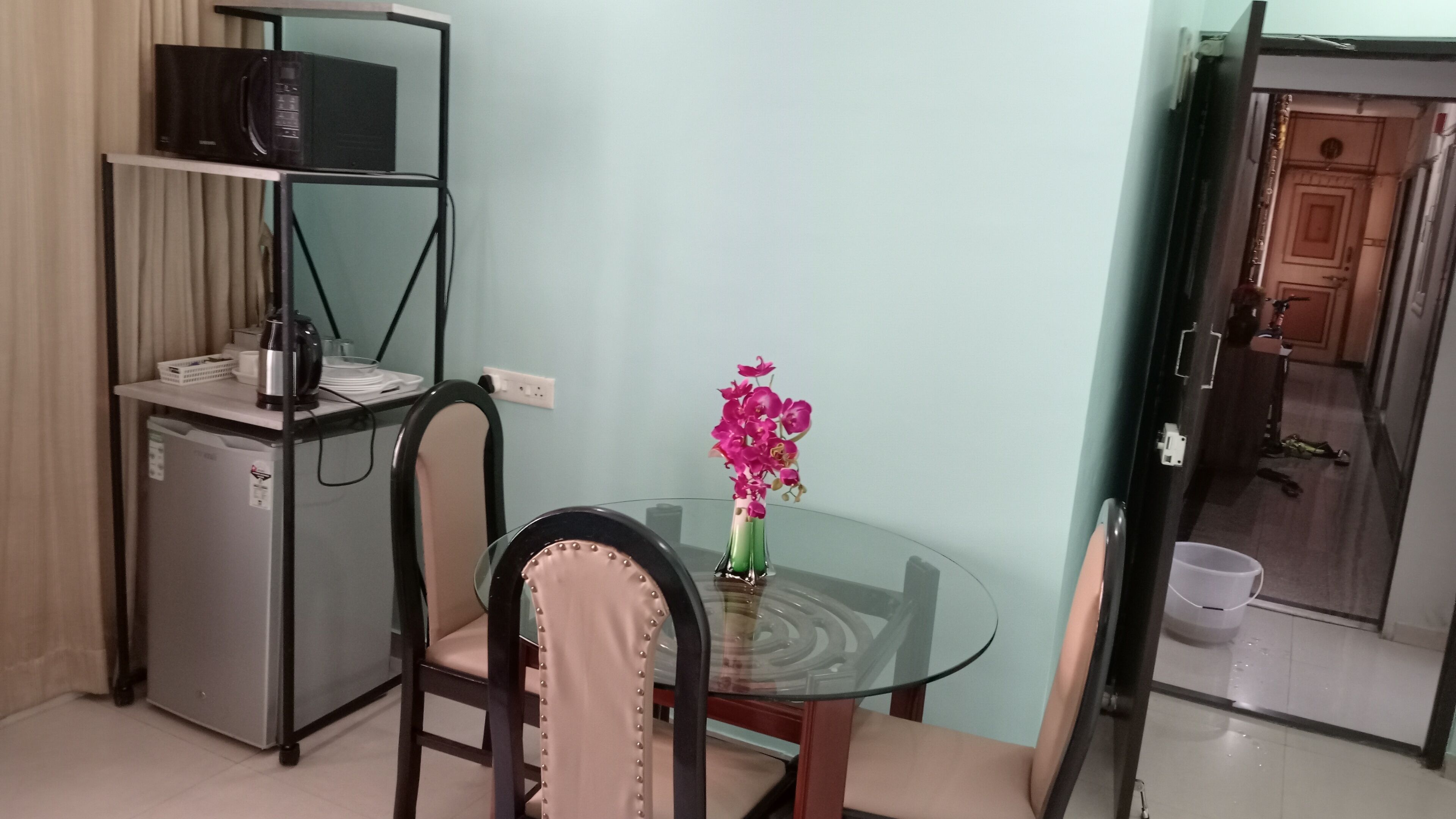 A compact room with a glass table, a chair, a small refrigerator, a microwave, a vase with flowers, and a door leading to a bathroom.