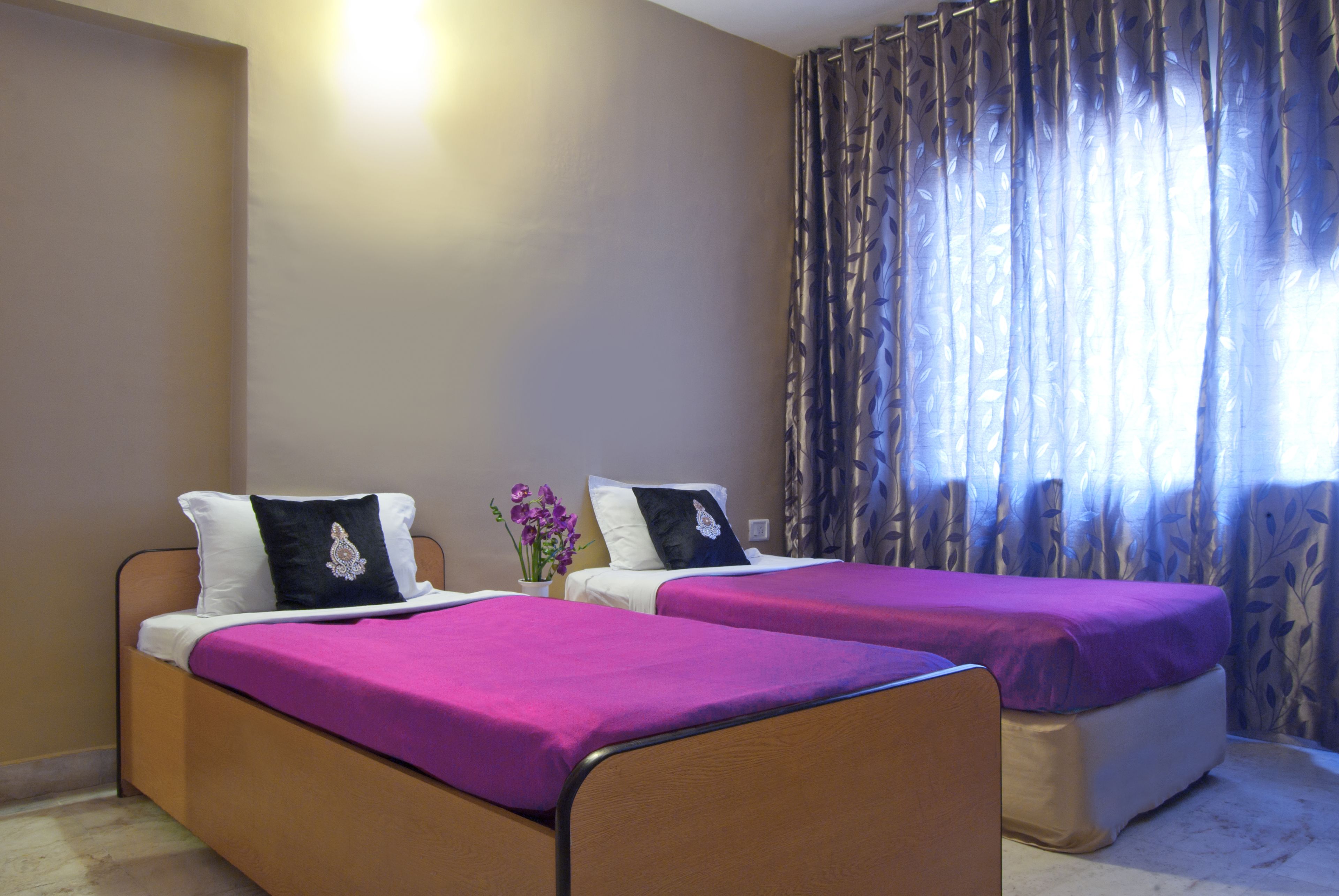 A room with two beds, each with a purple bedspread and white pillows. There is a vase with flowers on a nightstand between the beds.