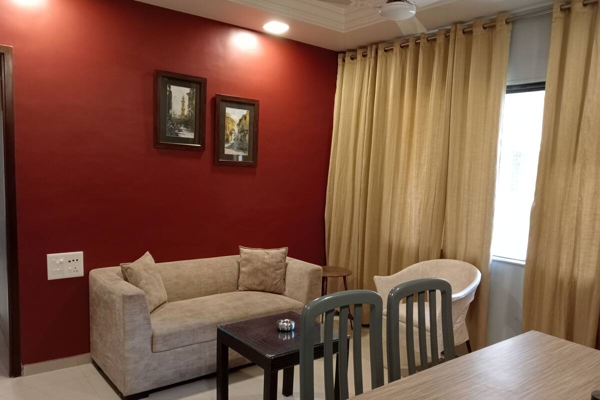 A room with a red wall, two framed pictures, a beige sofa, a small table, and a dining table with chairs.