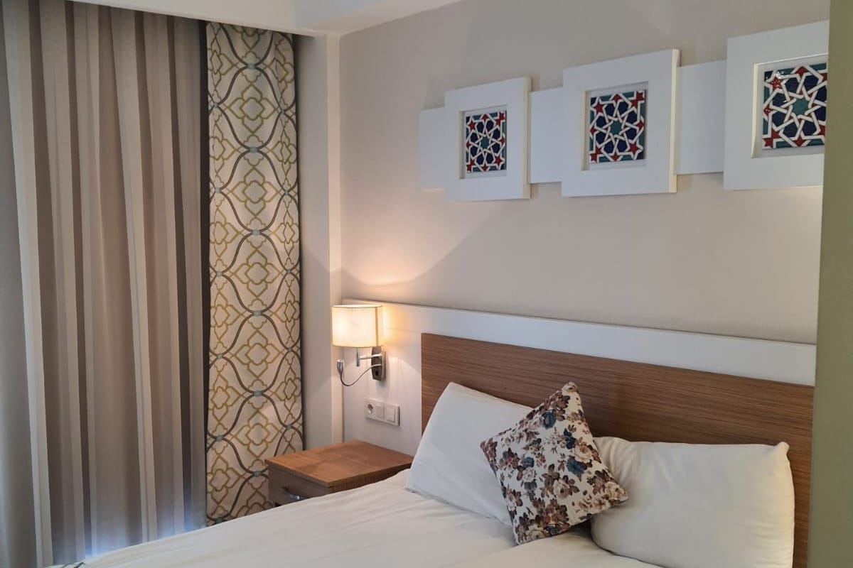 Una habitación de hotel con cama, mesita de noche, lámpara de pared y ventana con cortinas.