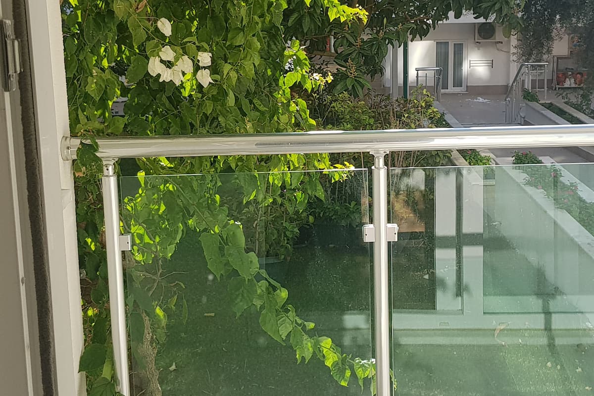 A balcony with a glass railing, surrounded by lush green foliage and flowering plants.