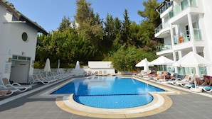 Piscina con sillones de descanso y sombrillas, rodeada de árboles y un edificio con balcones.