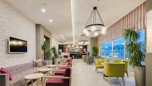 Un moderno vestíbulo de hotel con pared de ladrillo, un televisor de pantalla plana, varias zonas de estar con mesas y sillas redondas, y amplias ventanas con vistas a la ciudad.
