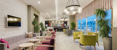 A modern hotel lobby with a brick wall, a flat-screen TV, multiple seating areas with round tables and chairs, and large windows offering a view of the city.