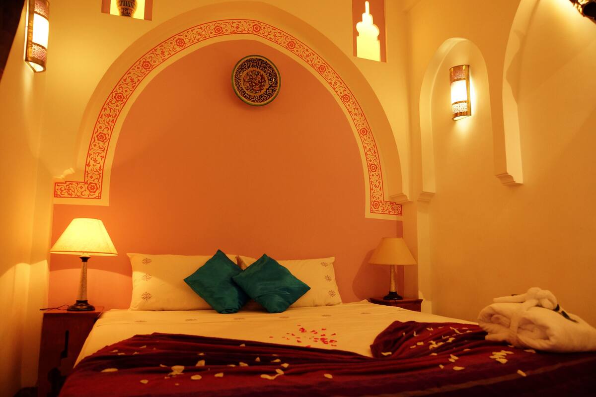 Habitación de hotel con cama, dos almohadas, una colcha, mesita de noche, lámpara y pared decorada con un arco y un aplique.