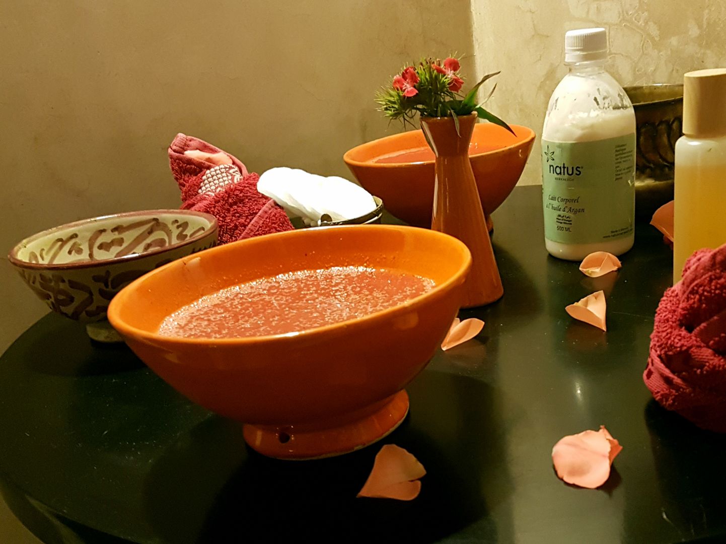 Un baño con un bol grande anaranjado, un bol más pequeño, una botella de loción, un florero con flores y una toalla roja.