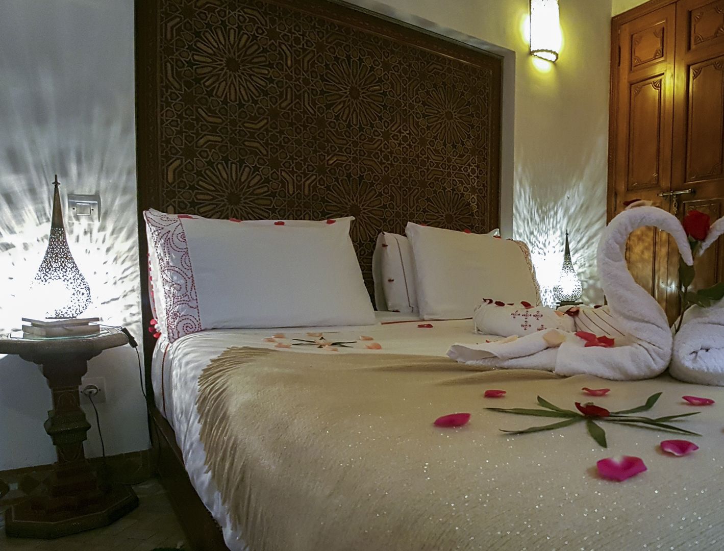 A hotel room with a bed, decorative pillows, a headboard with intricate patterns, a nightstand with a lamp, and a wooden wardrobe.