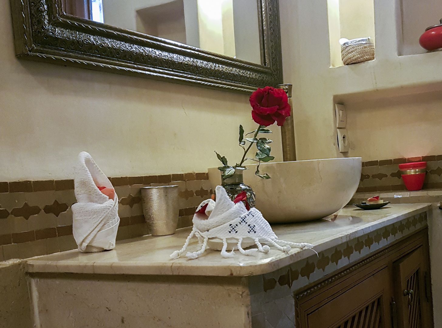 A bathroom with a large mirror, a bowl, a vase with a red rose, and towels on the counter.