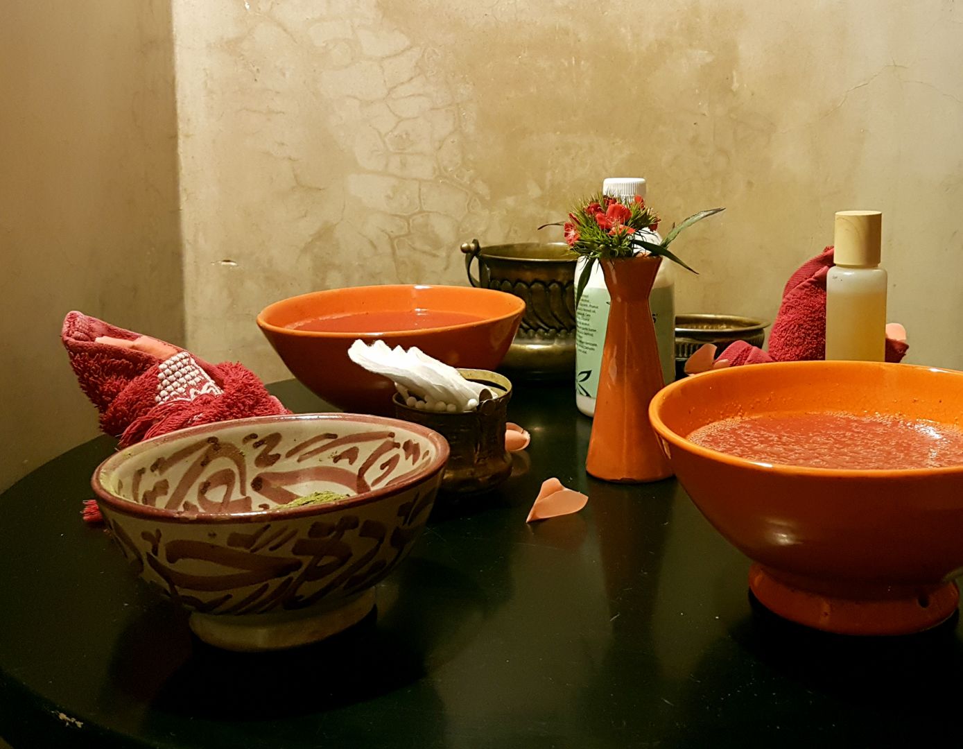 Un baño con encimera negra, un tazón naranja grande, un tazón más pequeño con motivos florales, un jarrón blanco con flores y una botella de loción.