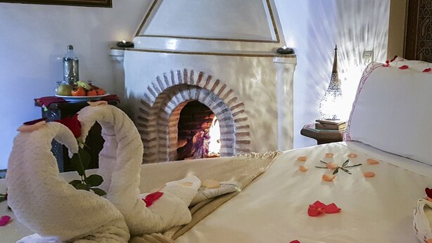 Una cama adornada con pétalos de rosa y una decoración de cisne, chimenea y una mesa con fruta y una botella.
