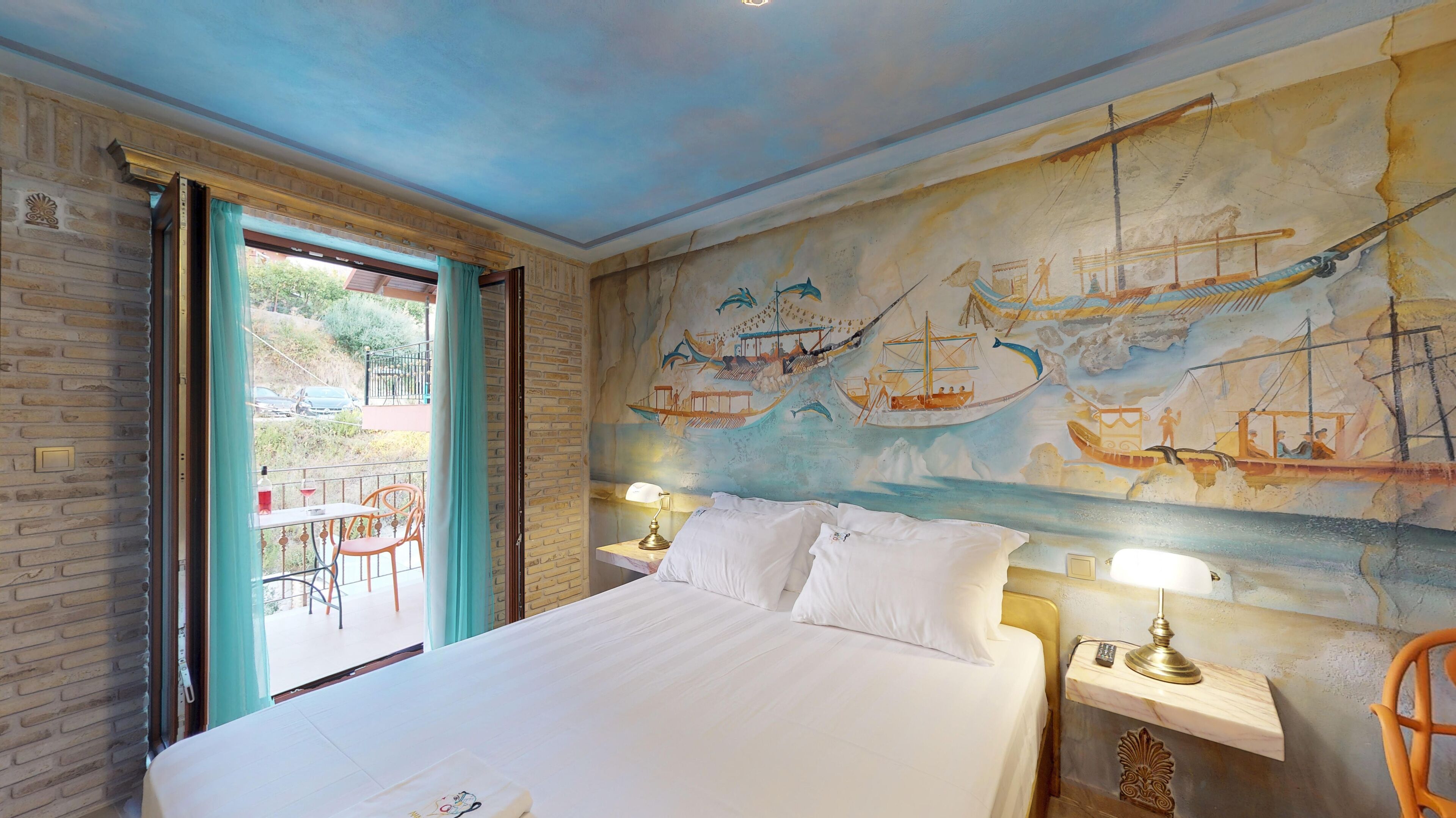Une chambre d’hôtel avec un grand lit, deux tables de chevet avec des lampes, une fresque représentant des navires sur le mur et un balcon avec des chaises.