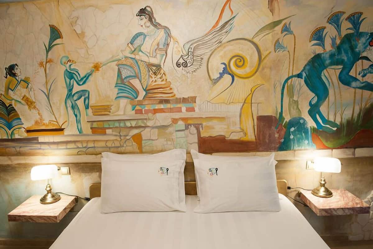 A hotel room with a mural depicting ancient Egyptian figures and a bed with white linens and matching pillows.