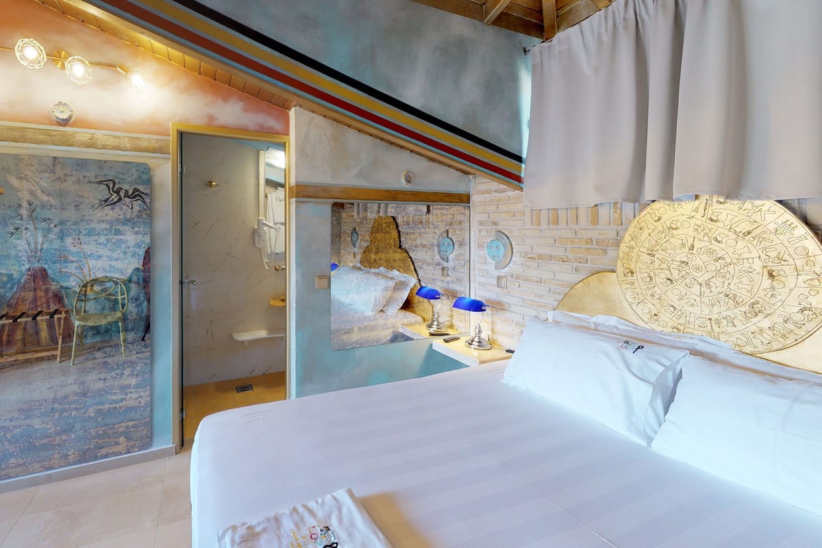 A hotel room with a bed, a desk with a lamp, a bathroom with a shower, and a mural on the wall.