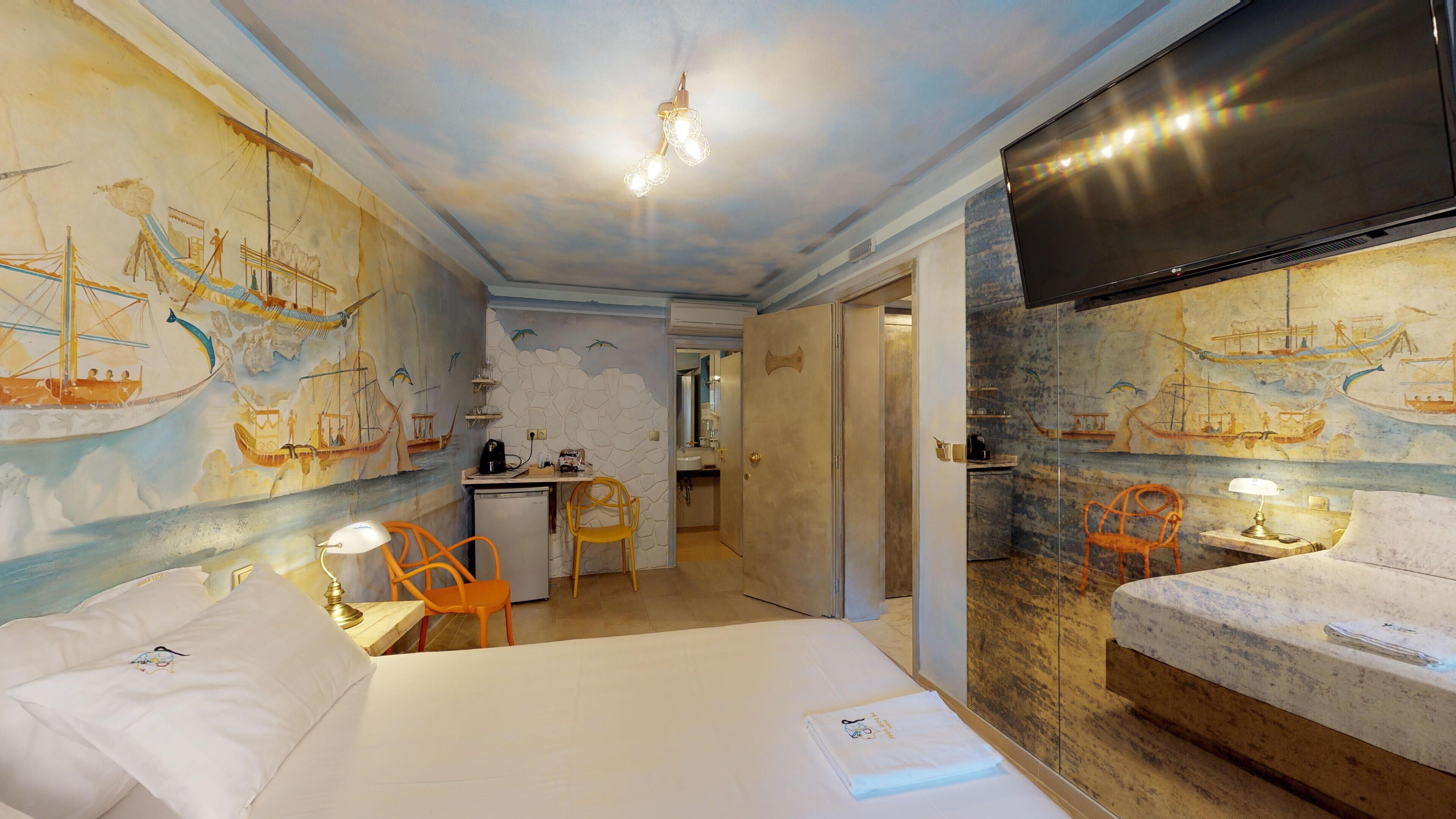 A hotel room with a bed, a desk, and a chair. There is a TV mounted on the wall and a mural depicting ships on the walls.