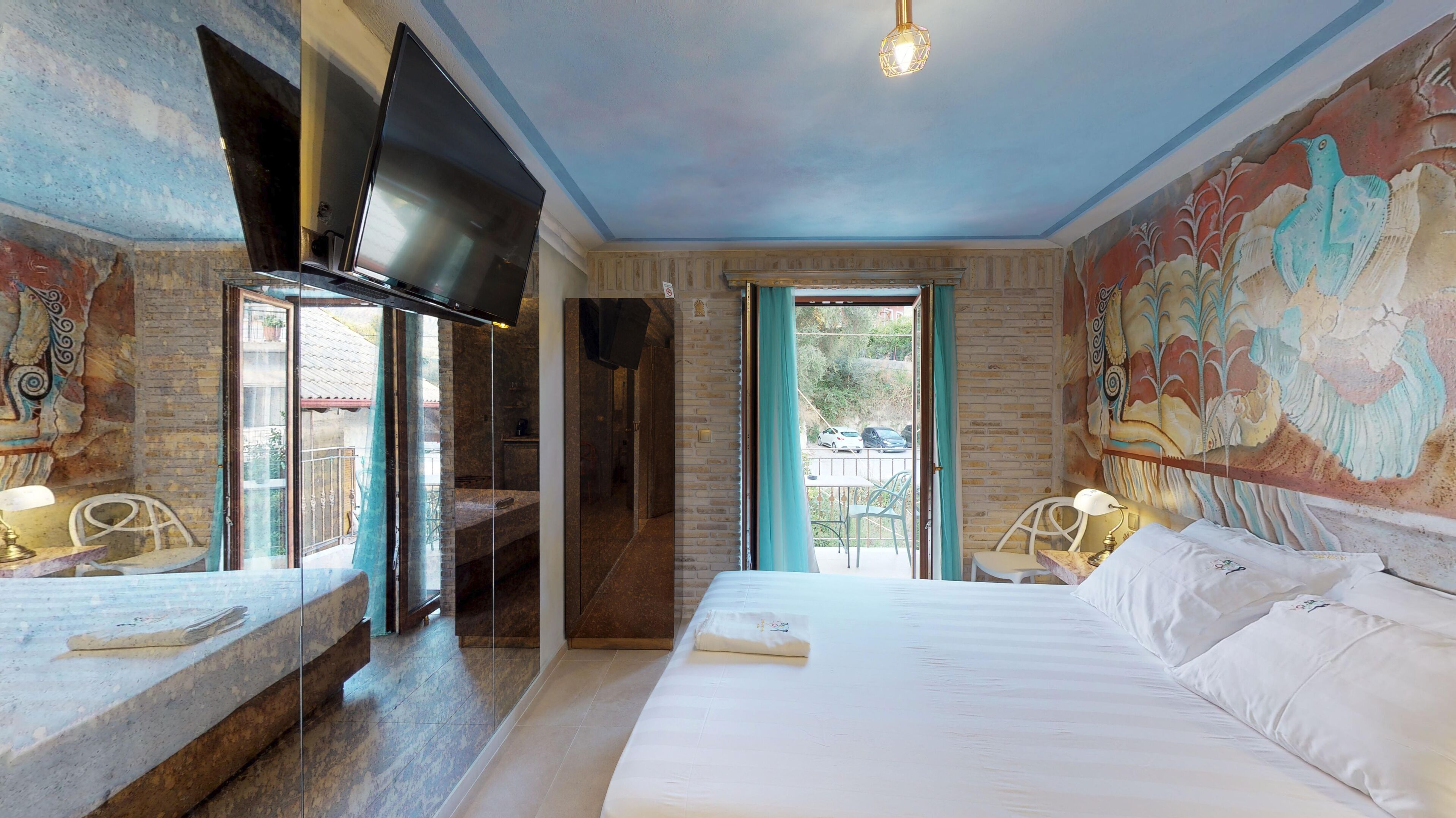 A hotel room with a large bed, a TV mounted on the wall, a balcony with a view, and a mural on the wall.