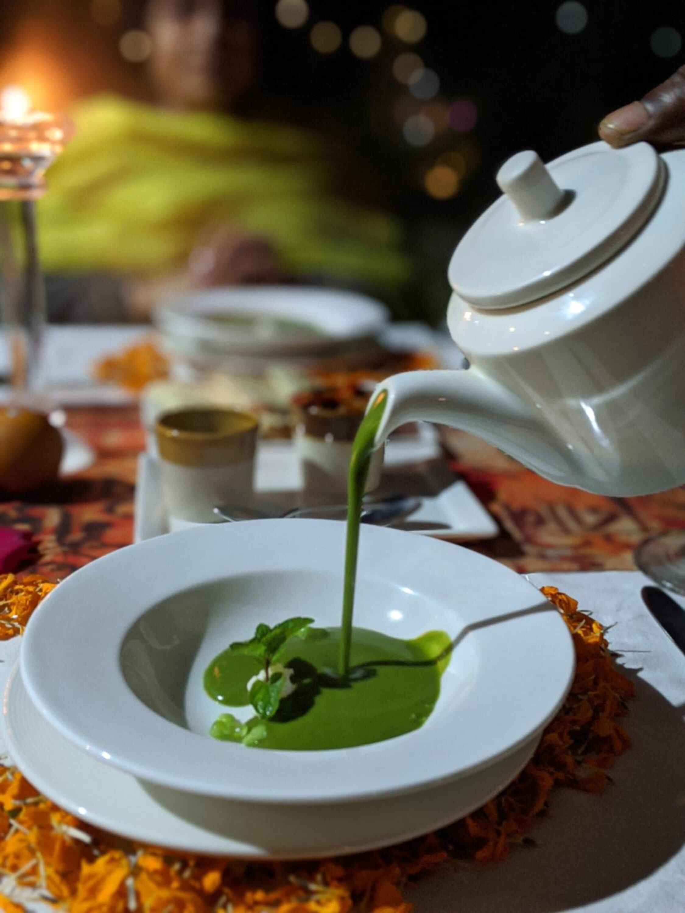 A white teapot pouring green liquid into a white bowl on a table with a candle in the background.
