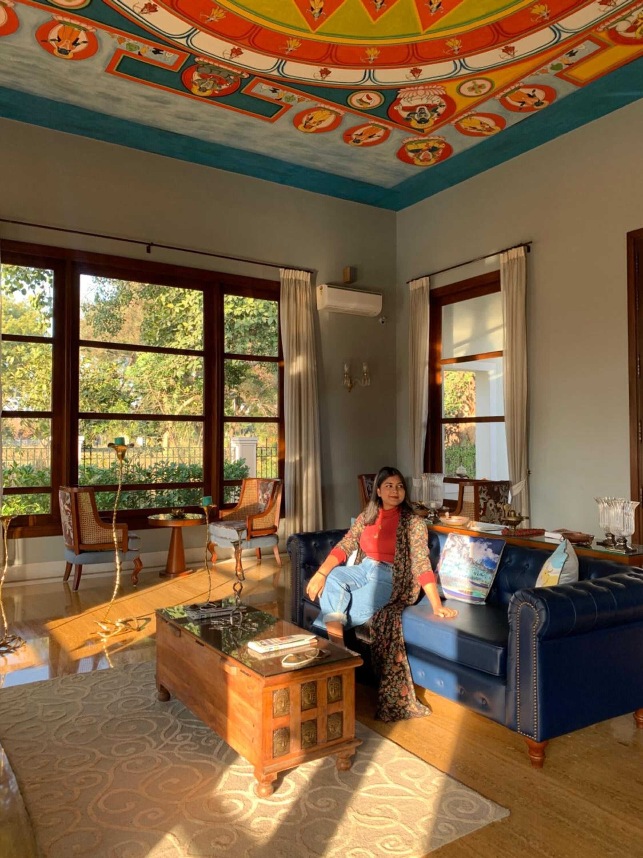 A living room with a blue sofa, a wooden coffee table, and a patterned rug. There are two windows with curtains, a ceiling with a colorful design, and a small table with a lamp.
