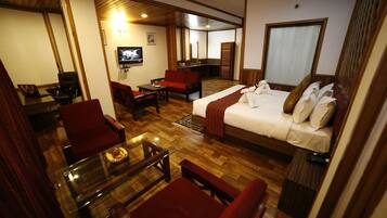 A hotel room with a bed, red chairs, a glass coffee table, a TV, and a wooden floor.