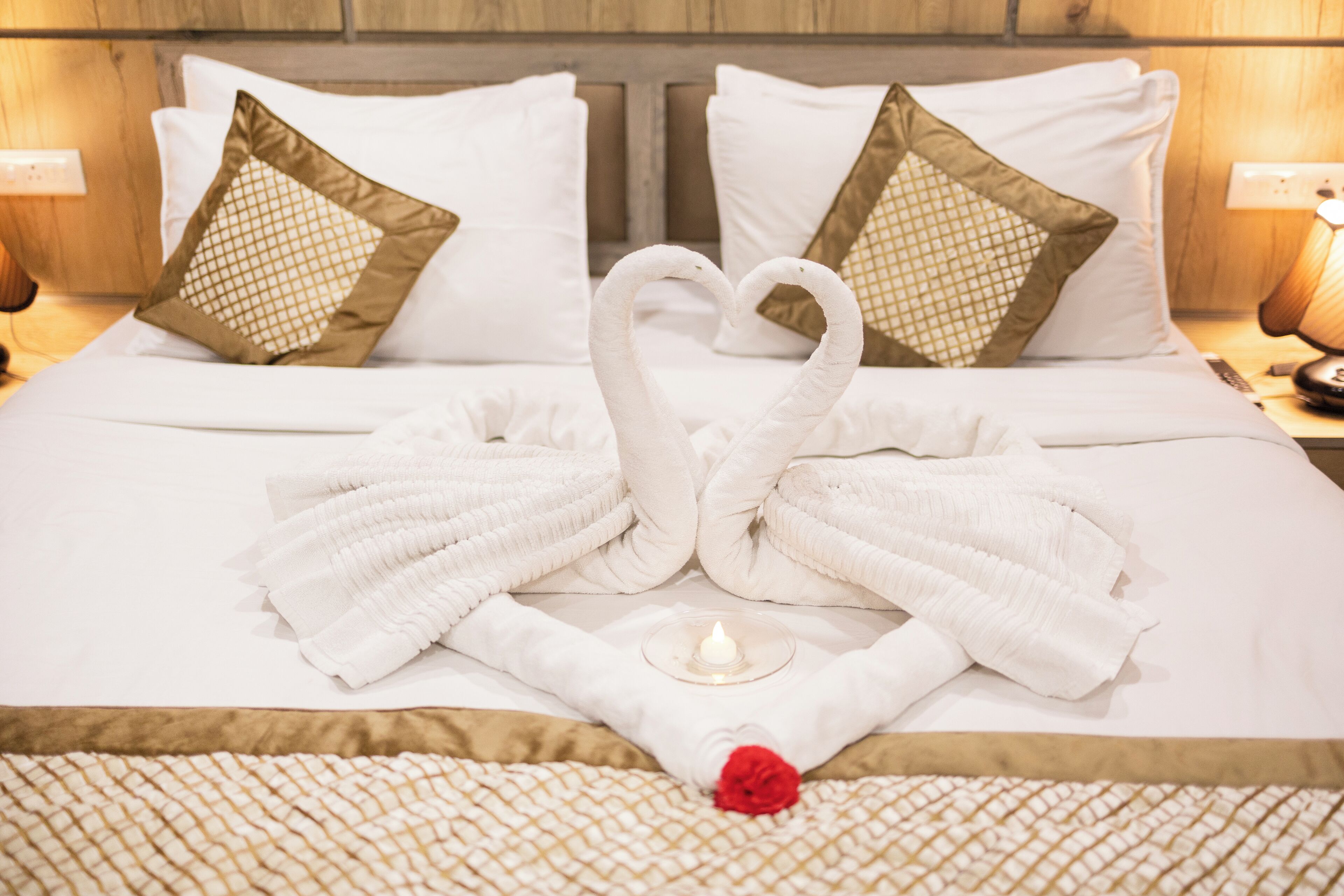 A neatly made bed with towels arranged in the shape of swans, a lit candle, and a red rose on a patterned bedspread.