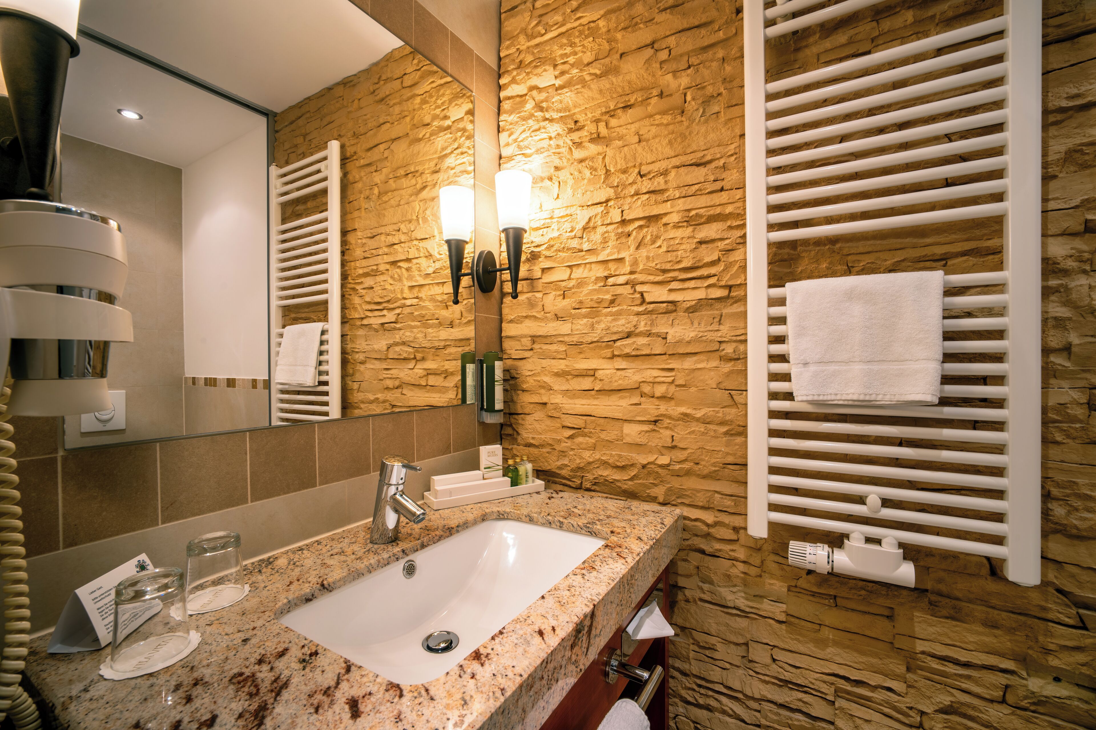 A bathroom with a granite countertop sink, a mirror, a towel rack, and a wall-mounted towel warmer.