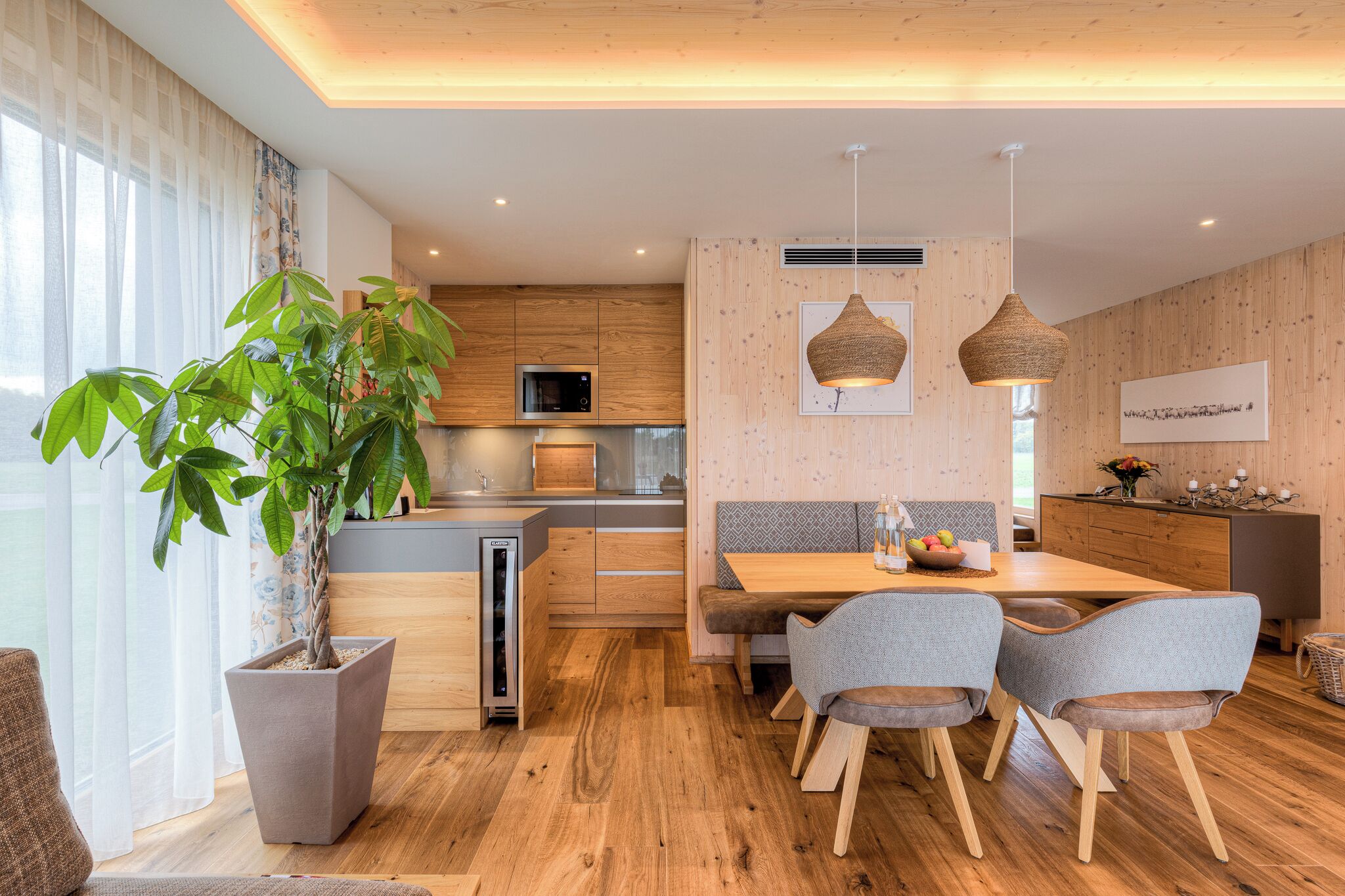 A modern kitchen with wooden cabinets, a built-in microwave, and a dining area with a wooden table and grey upholstered chairs.