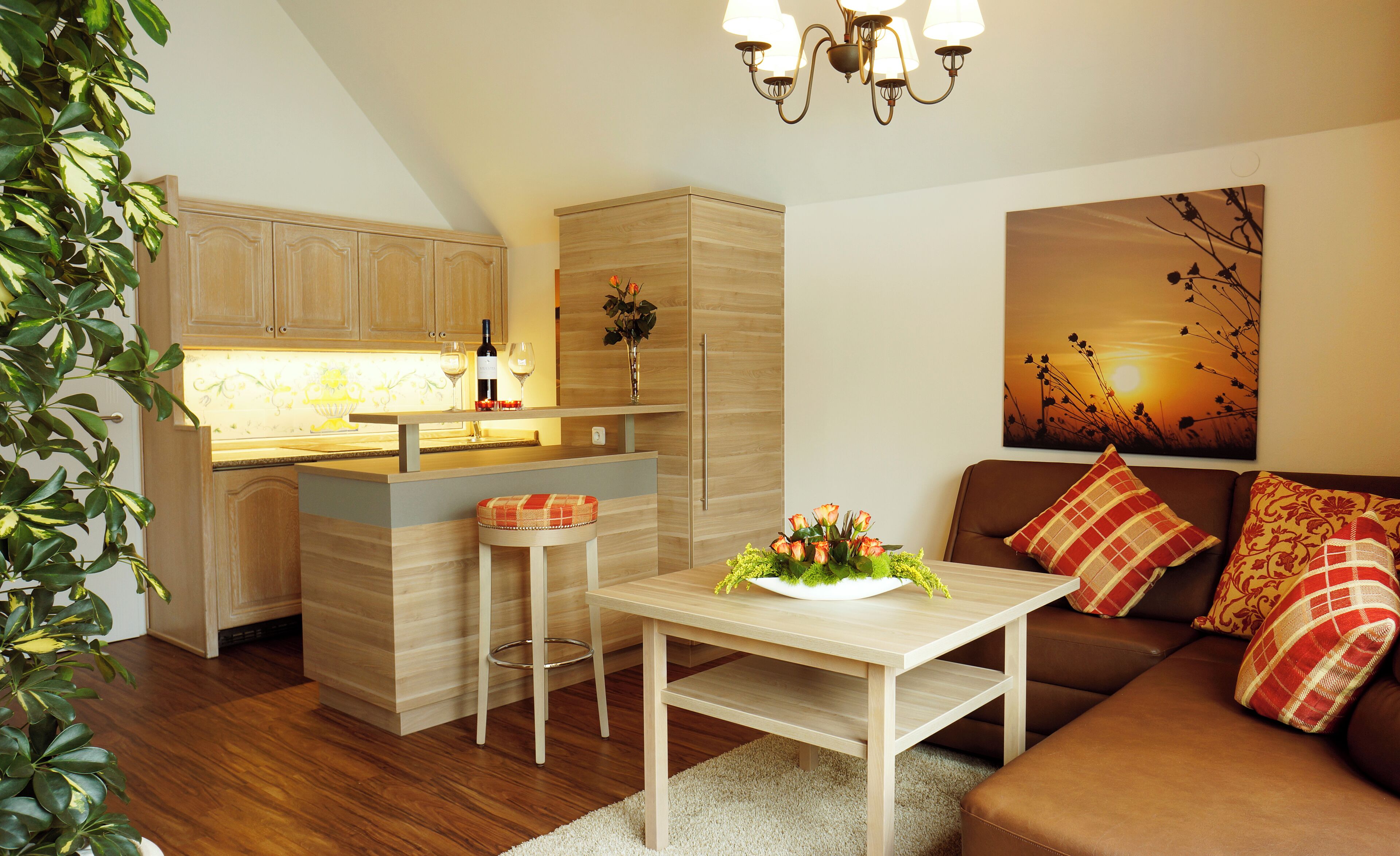 Eine moderne Küche mit Holzschränken, einer zentralen Kochinsel und einem Essbereich mit Tisch und Stühlen. Es gibt eine Couch mit gemusterten Kissen und ein Gemälde eines Sonnenuntergangs an der Wand.