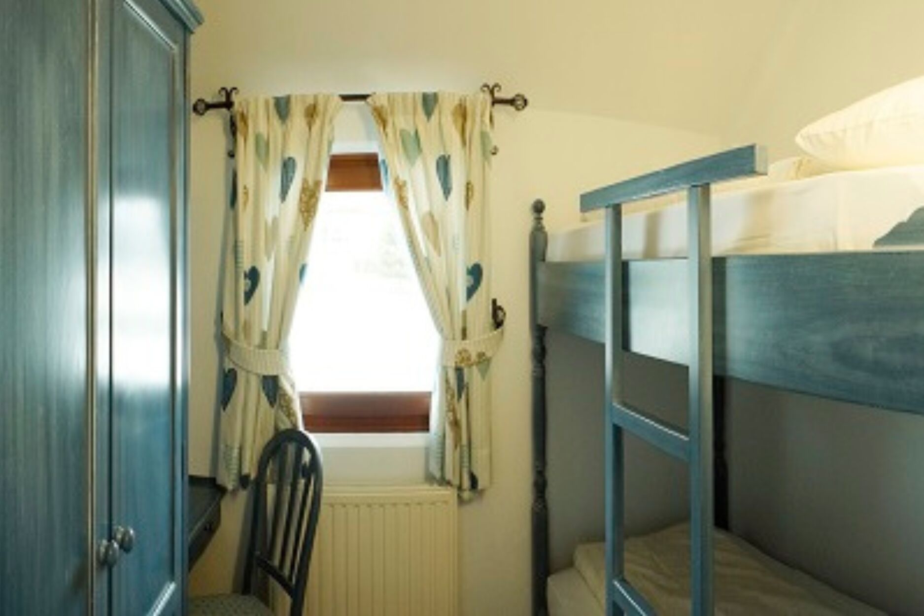 A room with a bunk bed, a chair, a radiator, and a window with curtains.