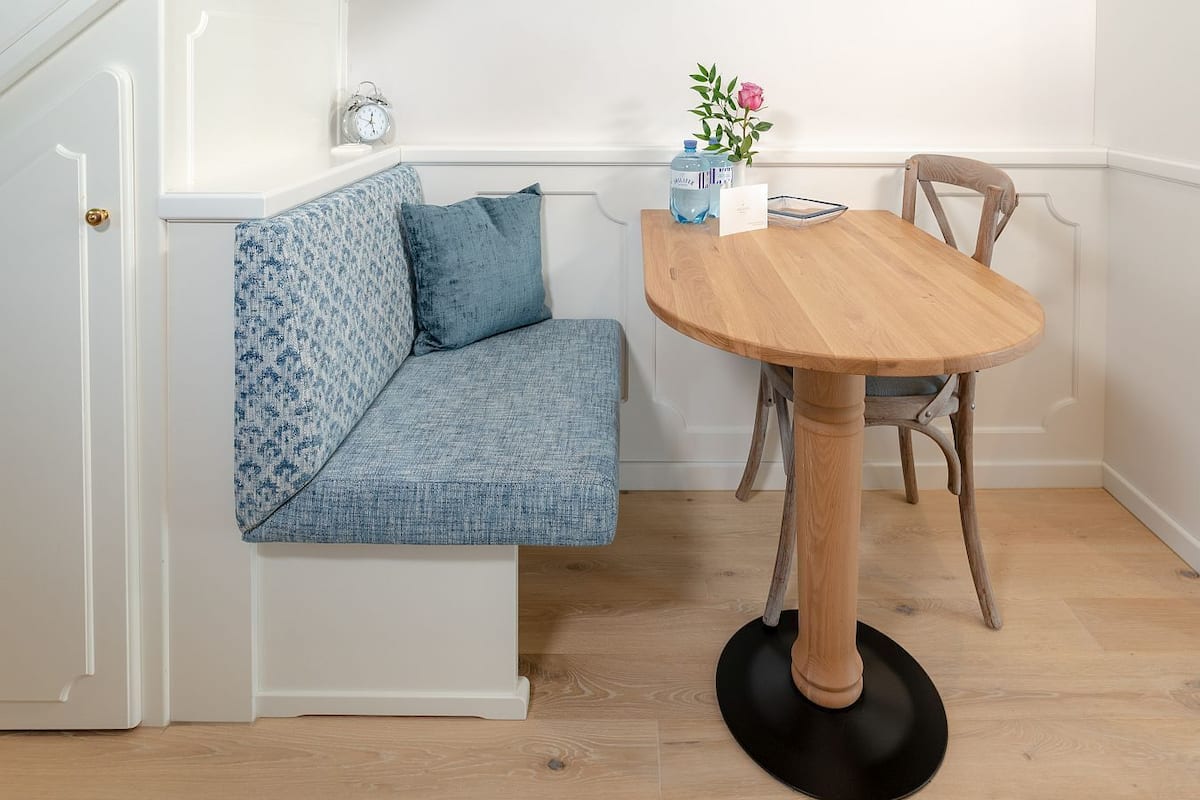 Een kamer met een hoekopstelling, een houten eettafel, een bank met blauwe kussens, een klok aan de muur en een vaas met bloemen op de tafel.