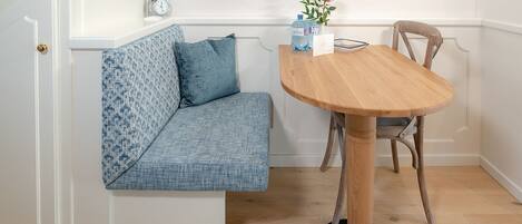 A corner room with a wooden dining table, a bench with blue cushions, a clock on the wall, and a vase with flowers on the table.