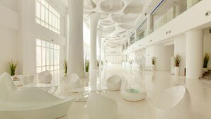 Een ruime lobby met modern wit meubilair, grote ramen en een hoog plafond met bijzondere architectonische details.