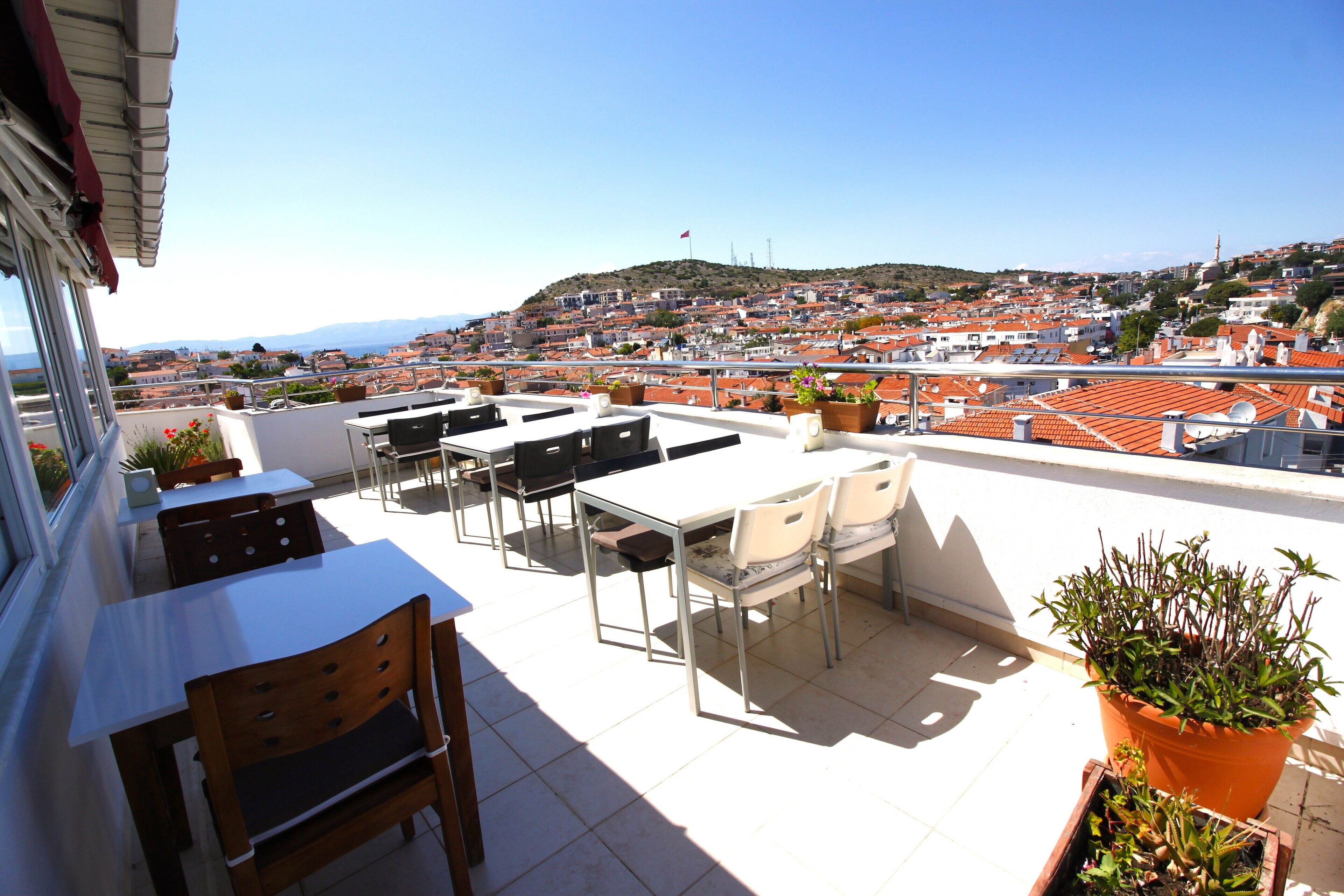 A rooftop terrace with tables and chairs, potted plants, and a view of a cityscape with red-roofed buildings.