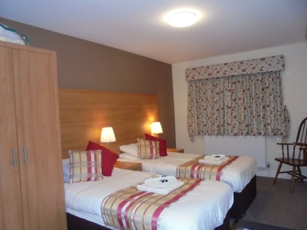 A hotel room with two beds, wooden headboard, bedside lamps, floral curtains, and a wooden chair.