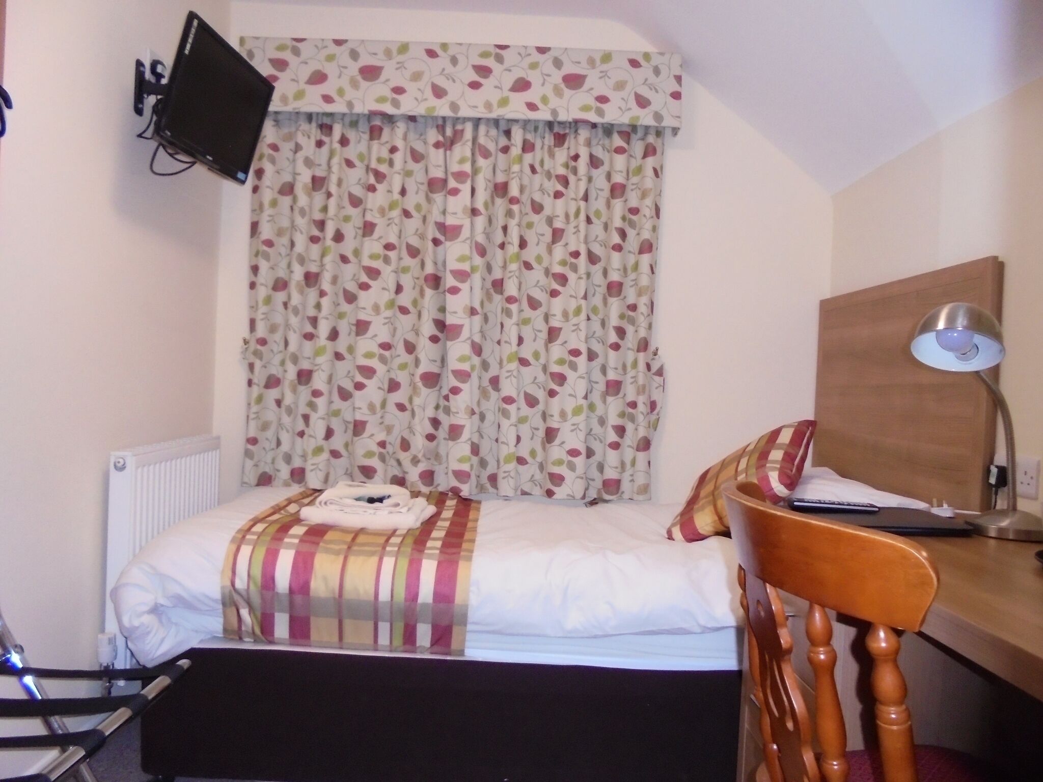 Ein kleiner, ordentlicher Raum mit einem Bett, einem Schreibtisch, einem Stuhl, einer Schreibtischlampe und einem an der Wand befestigten Fernseher.