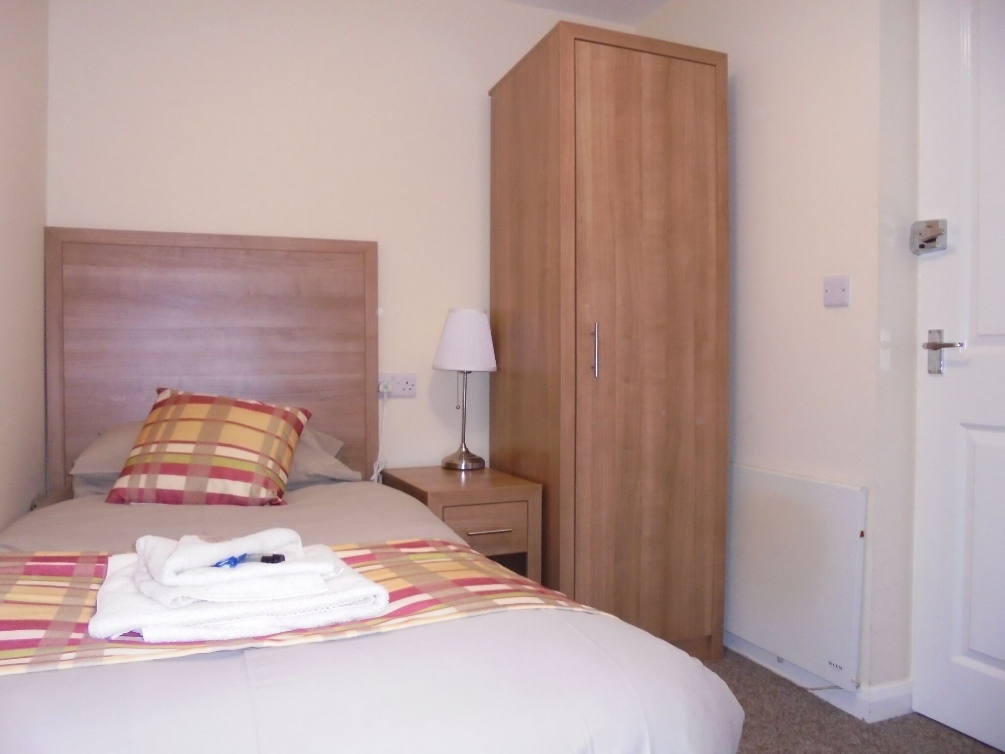 A hotel room with a bed, a wooden wardrobe, a nightstand with a lamp, and a radiator.
