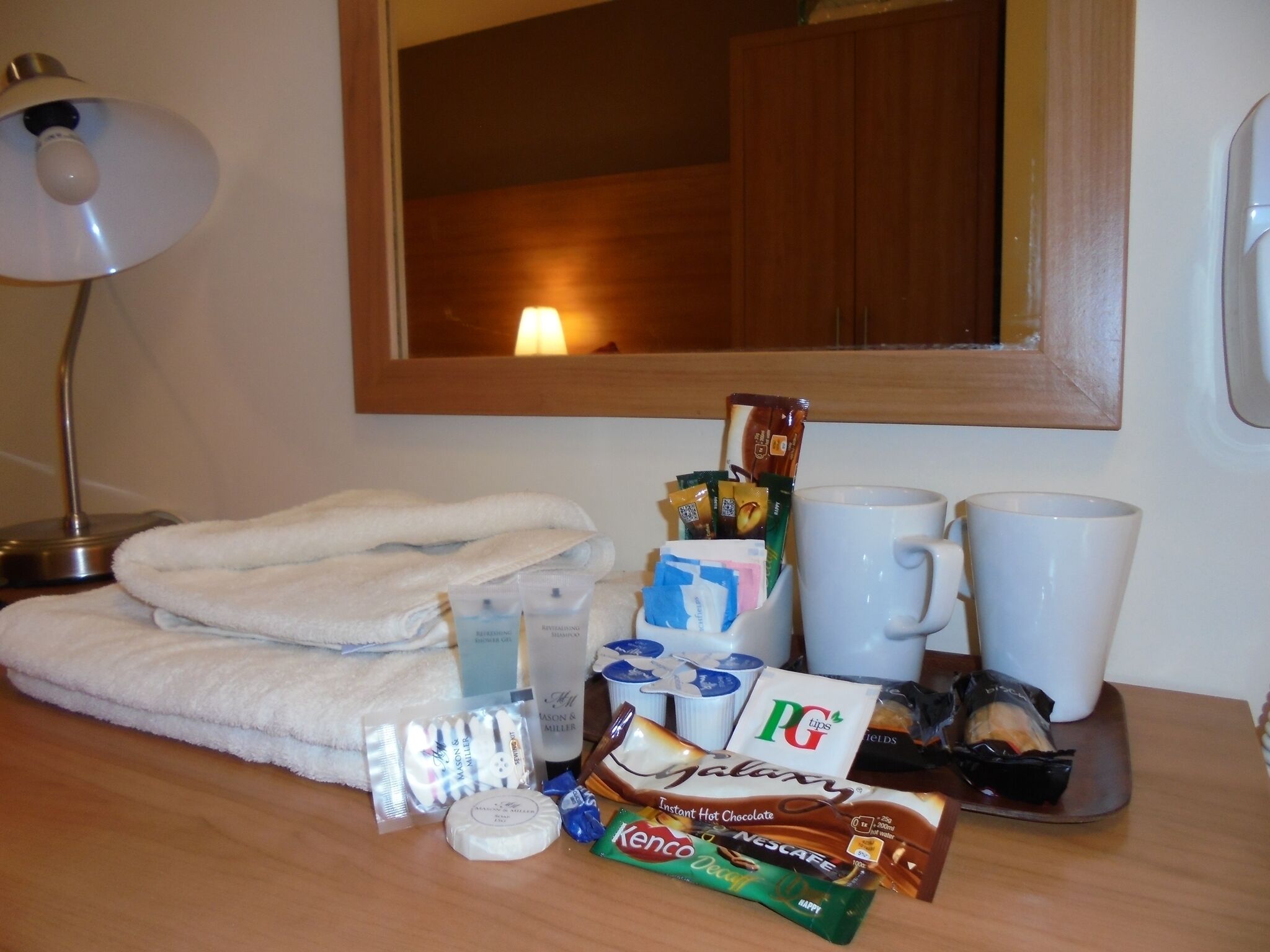 Habitación de hotel con una toalla blanca doblada con cuidado, una lámpara de noche, una taza de café instantáneo, una mug, una bandeja con aperitivos y artículos de aseo.