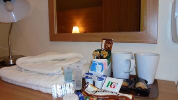 Habitación de hotel con una toalla blanca doblada con cuidado, una lámpara de noche, una taza de café instantáneo, una mug, una bandeja con aperitivos y artículos de aseo.