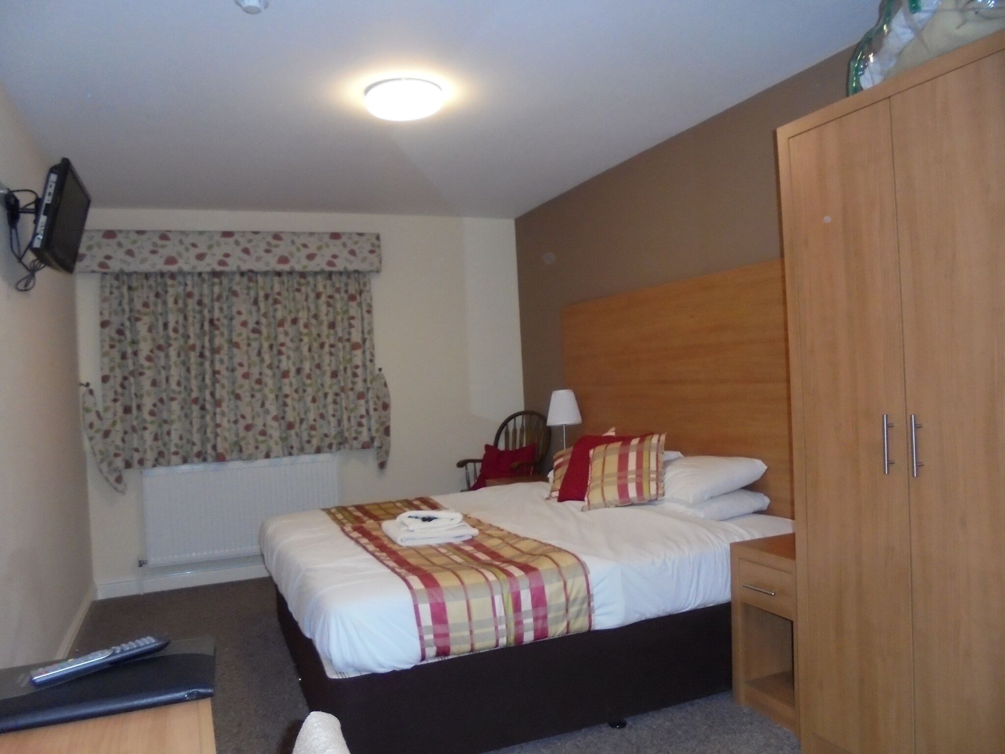 A hotel room with a bed, a wooden wardrobe, a desk with a remote control, a lamp, and a window with floral curtains.