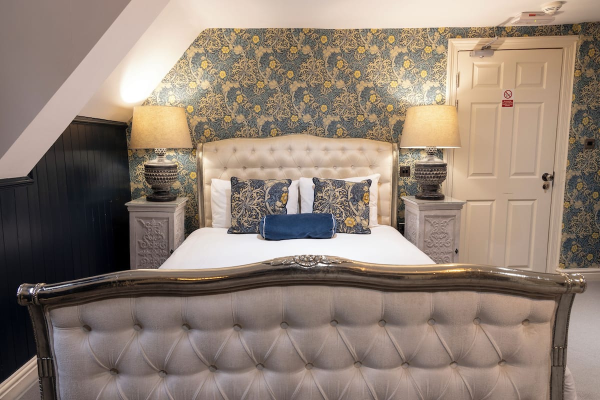 A bedroom with a tufted headboard, a bed with white bedding, two bedside tables with lamps, and floral wallpaper.