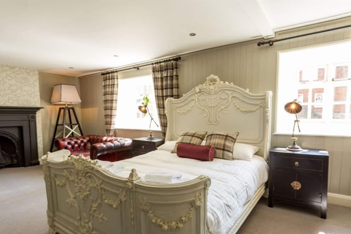 A bedroom with a large bed, a fireplace, a leather armchair, and a nightstand with a lamp.