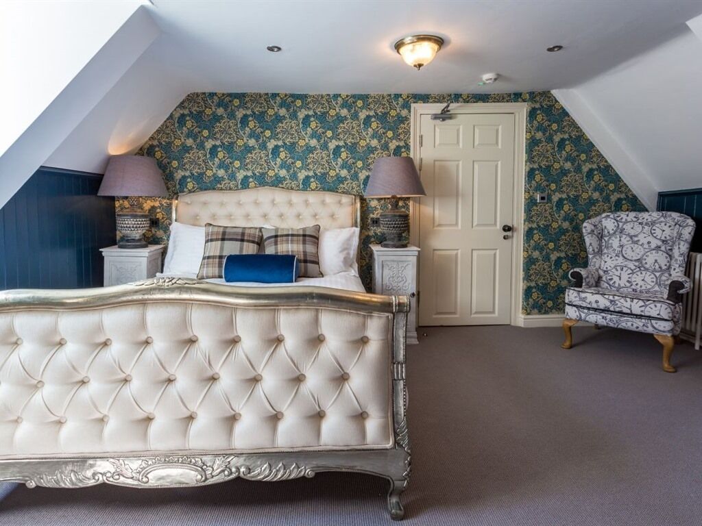 A bedroom with a tufted bed, two bedside tables with lamps, a door, and a patterned armchair.