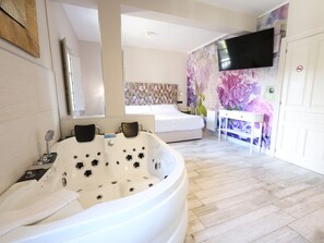 Een moderne badkamer met een groot bad, een bed met een bloemenbekleding bij het hoofdeinde, en een televisie aan de muur bevestigd.