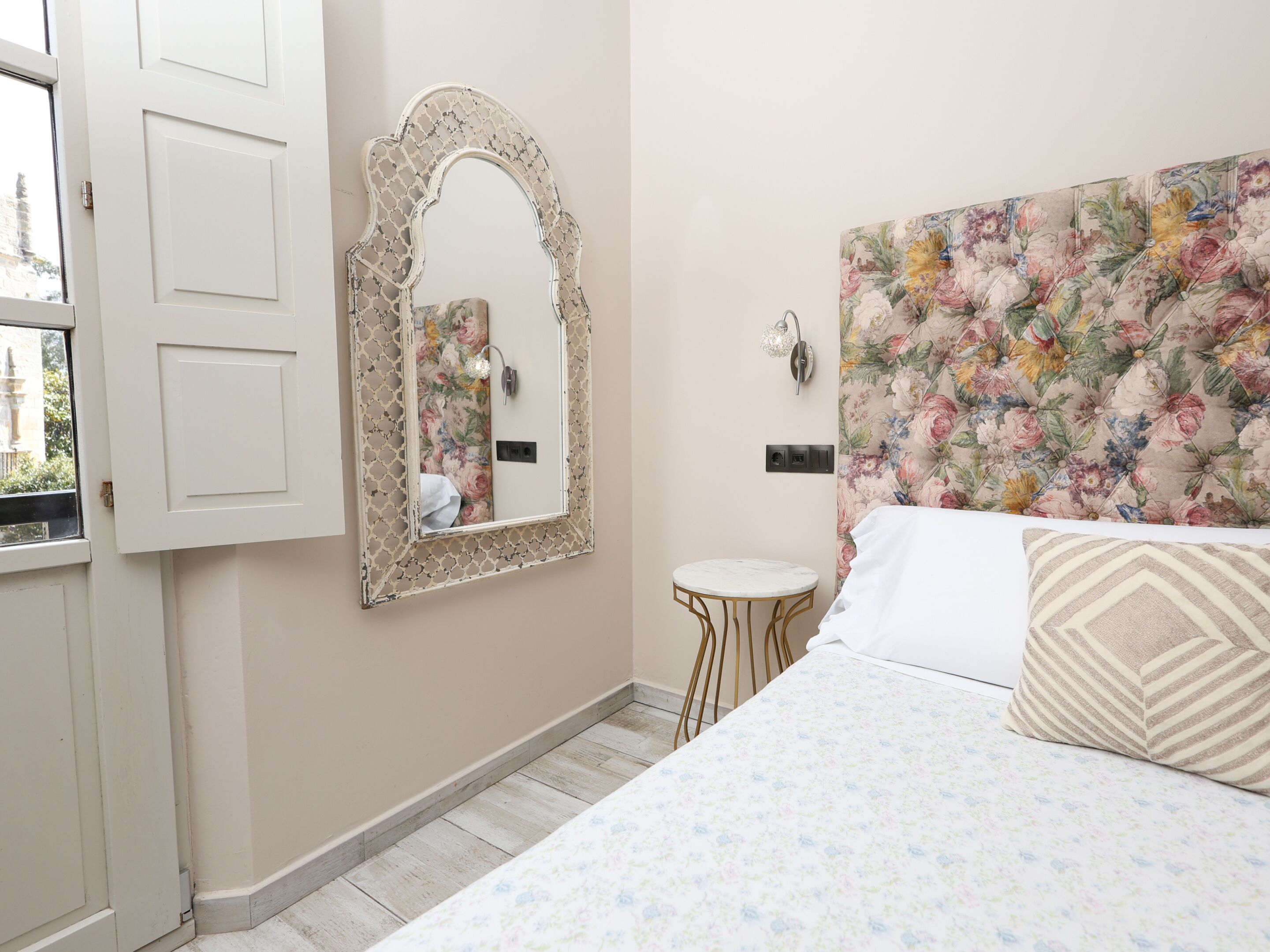 A bedroom with a bed, a mirror, a small round table, and a floral headboard.