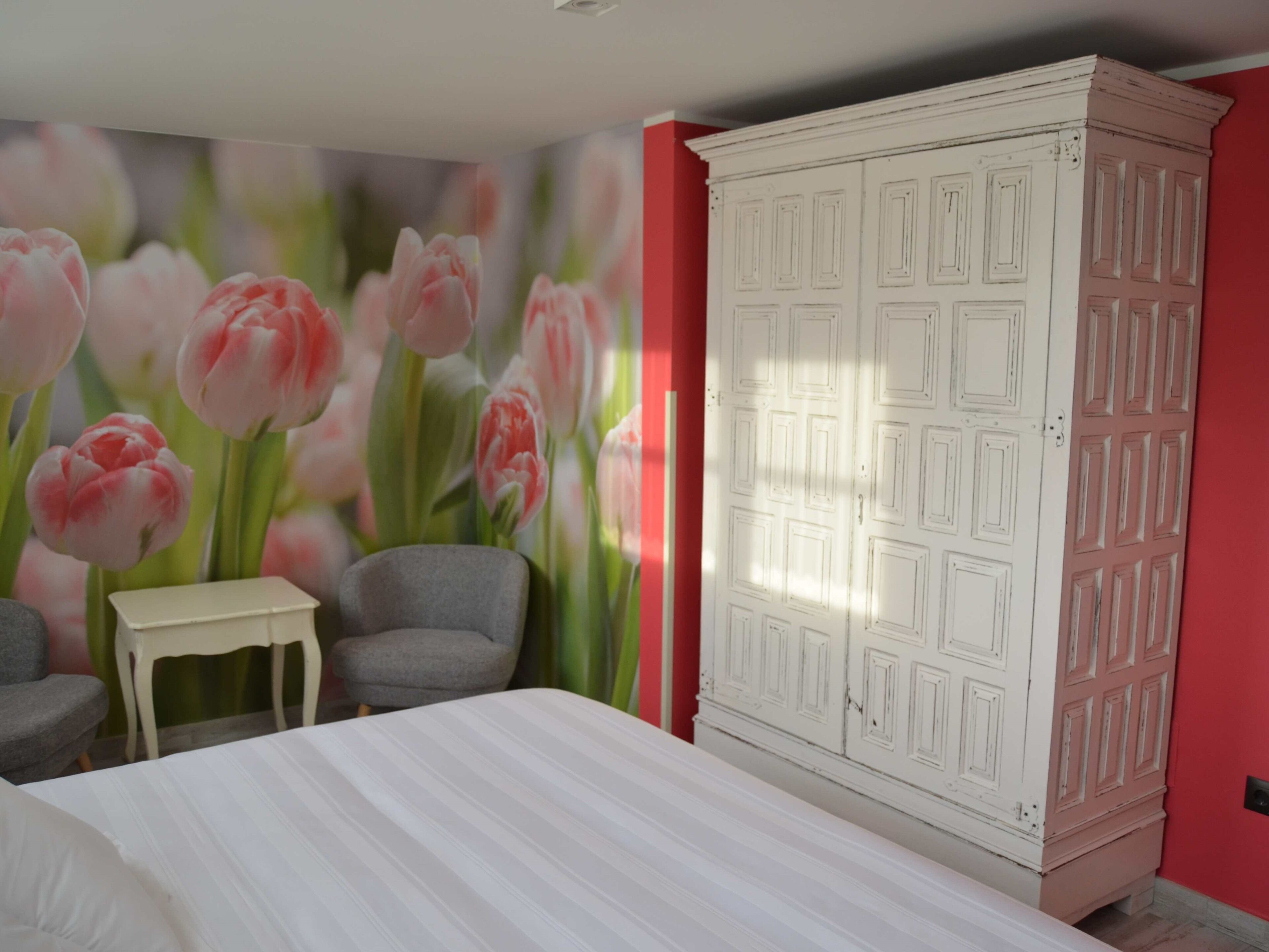 Een slaapkamer met een bed, een kledingkast, twee stoelen, een kleine tafel en een muur met een bloemmotief.