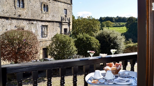 Un balcón con una mesa lista para el desayuno, con vista a un edificio de piedra histórico y un paisaje campestre.
