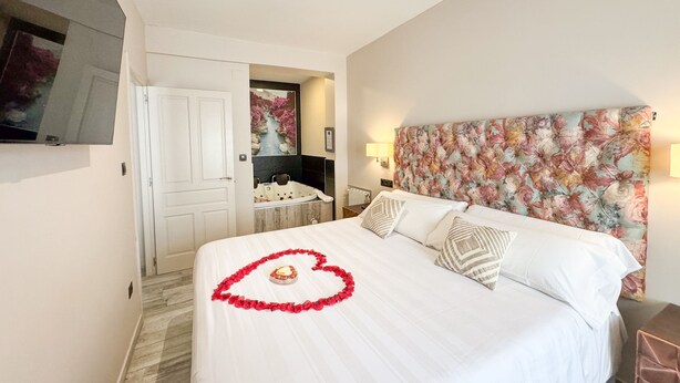 A hotel room with a bed, a television, a bathroom with a bathtub, and a floral headboard. - Akla Hotel Suites Santillana (Santillana del Mar)